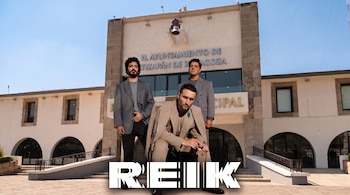 Tres hombres con trajes formales posan frente a un edificio blanco con arcos y campana, con la palabra Reik en letras blancas al frente.