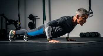 Hombre de cabello canoso en posición de flexión sobre una colchoneta gris en un gimnasio; viste ropa deportiva gris y azul petróleo, con un reloj verde oliva. Al fondo, pesas.