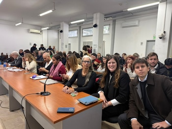 Vista de una sala de audiencia con varias personas sentadas en mesas largas y filas, algunas con laptops y documentos, durante un procedimiento judicial