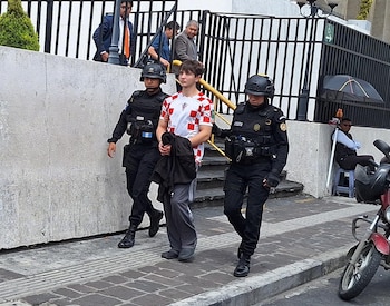 Dos agentes policiales con uniforme negro y casco escoltan a un joven con camiseta a cuadros rojos y blancos. Se observan un muro, una reja negra y una motocicleta