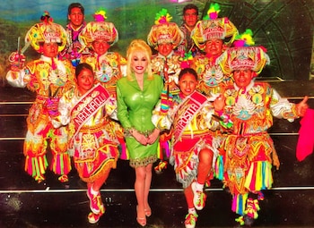 Dolly Parton con traje verde posa en el centro rodeada por integrantes de una agrupación de danzantes de tijeras con trajes coloridos