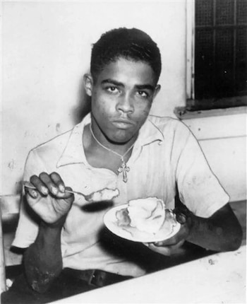 Fotografía en blanco y negro de un hombre joven sentado a una mesa con un plato de pastel, una cuchara y una cruz en el cuello