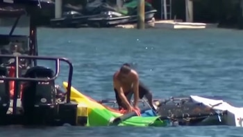 Un hombre sin camisa se inclina sobre un bote volcado, parcialmente sumergido en el agua. Se ven otros botes y un muelle en el fondo