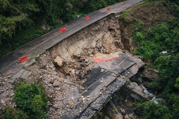 Carretera asfaltada con una sección central colapsada, rodeada de escombros, rocas y vegetación verde, con barreras de tráfico naranjas.