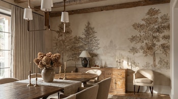 Comedor con mesa de madera, sillas tapizadas y un mural de árboles en la pared trasera. Un candelabro cuelga del techo con vigas expuestas.
