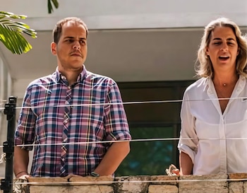 Magalli Meda, con cabello rubio y camisa blanca, y Pedro Urruchurtu, con camisa a cuadros, se asoman por un muro de ladrillos