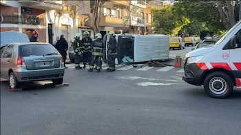 Una furgoneta volcada de costado en una calle junto a bomberos uniformados, un automóvil gris y una ambulancia en primer plano