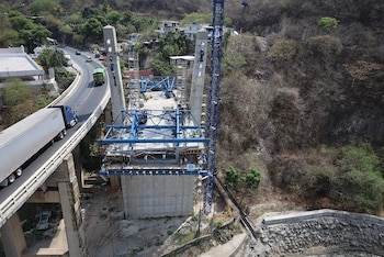 Una vista aérea de un puente en construcción con andamios azules, al lado de una carretera transitada por camiones, sobre un terreno montañoso con vegetación