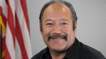 Hombre de camisa negra y bigote sonriendo ante la bandera de Estados Unidos