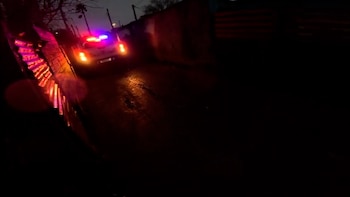 Vista trasera de un coche de policía con luces de emergencia encendidas en una calle oscura y húmeda. La luz roja y azul ilumina una pared