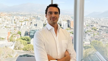 Hombre de camisa blanca con los brazos cruzados ante un ventanal que da a una ciudad y montañas de fondo