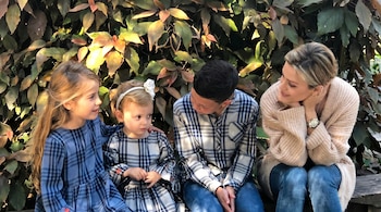 Una mujer sonriente y tres niños, dos niñas y un niño, sentados en un banco de madera frente a un fondo de hojas verdes y rojizas bajo la luz del sol.
