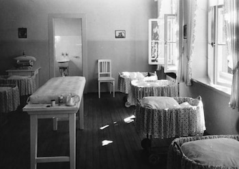 Habitación en blanco y negro con varias cunas, una mesa larga con colchón, una silla y ventanas con cortinas. Un lavabo se ve a través de una puerta