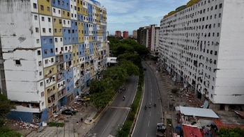 Dos edificios residenciales altos con fachadas deterioradas flanquean una calle. Escombros en la base de los edificios. Árboles y vehículos circulan por la calle