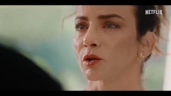 Primer plano de una mujer con cabello oscuro, ojos claros, labios pintados de rojo y un pendiente dorado en la oreja derecha con forma de sol. Logo de Netflix arriba a la derecha