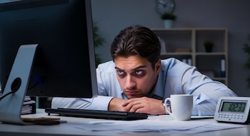 Un hombre con ojeras pronunciadas se apoya en un escritorio oscuro frente a un monitor de computadora, con una taza de café y un reloj digital a su lado.