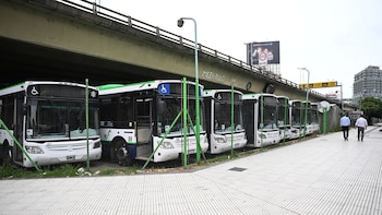 Autobuses blancos y verdes estacionados en fila bajo un puente de hormigón, detrás de una valla verde. Dos hombres caminan por la acera a la derecha