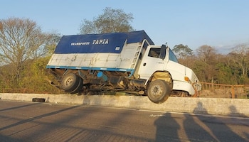 Camión blanco con lona azul de Transporte Tapia inclinado y montado sobre una barrera de hormigón en la carretera