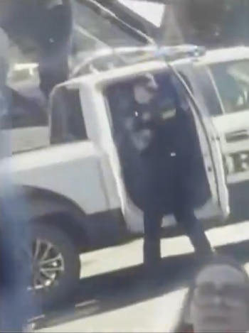 Un agente de policía de pie junto a una camioneta blanca con la puerta abierta en una calle