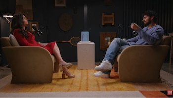 Un hombre y una mujer sentados en sillones frente a frente en un estudio de podcast, con un libro azul en un pedestal y micrófonos visibles