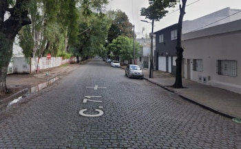 Vista diurna de calle adoquinada con vehículos estacionados, árboles, casas, un muro con grafiti y un cartel de prohibido el paso