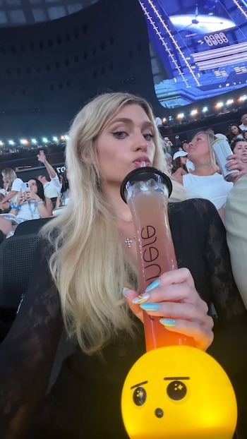 Mujer rubia bebiendo de un vaso grande con sorbete. Exhibe uñas celestes y amarillas, collar con cruz y un objeto luminoso amarillo. Personas en segundo plano