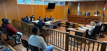 Jueces, abogados y acusados permanecen sentados en una sala de audiencias con madera y la bandera de Argentina al fondo