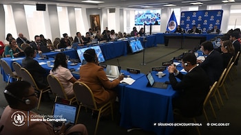 Personas sentadas ante mesas azules con computadoras portátiles, micrófonos y carteles con nombres de países en un salón de conferencias