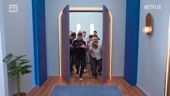 Nueve hombres jóvenes corren sonriendo por una puerta abierta hacia un pasillo, en un set de televisión con paredes azules y piso de madera