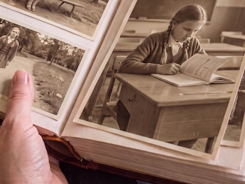 Manos sostienen un álbum de fotos abierto con tres imágenes sepia. Una niña está sentada en un banco, otra niña camina, y una niña lee un libro en un pupitre.