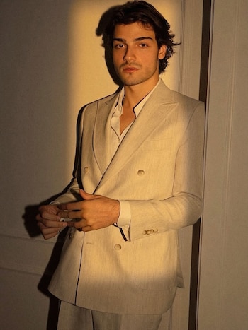 Un hombre joven de pelo oscuro y barba posa con un traje de lino beige, apoyado en una pared, bajo una luz cálida lateral que crea sombras