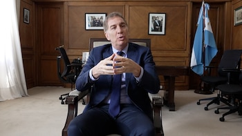 Hombre en traje azul sentado en un sillón club oscuro, gesticulando con las manos frente a un escritorio de madera. Una bandera argentina y sillas de oficina se ven al fondo
