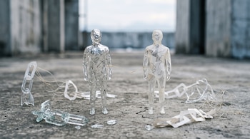 Dos esculturas transparentes con forma humana están unidas por un hilo fino sobre un suelo de concreto rodeadas de eslabones rotos.