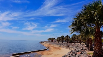 Una playa de arena con palmeras en la orilla, rocas apiladas a lo largo de la costa y agua tranquila bajo un cielo azul con nubes tenues.