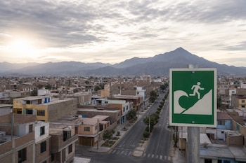 Cartel verde con pictograma de evacuación ante una ola, frente a una calle vacía, viviendas bajas y montañas al fondo.
