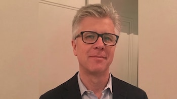 Hombre con cabello canoso, gafas rectangulares de montura oscura, chaqueta oscura, camisa azul clara con botones desabrochados, frente a una pared de tono claro.