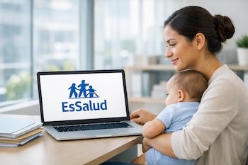 Mujer con bebé en brazos sentada frente a una laptop con el logo de EsSalud en la pantalla, documentos apilados en escritorio.