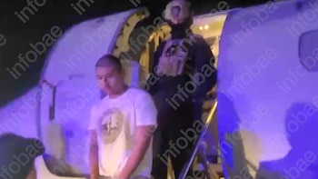 Un hombre en camiseta blanca es escoltado por una persona con equipo táctico en la rampa de un avión por la noche, bajo iluminación azul