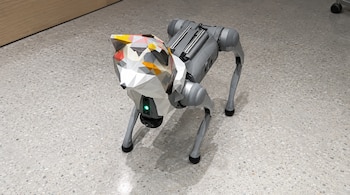 Robot cuadrúpedo de color gris con una máscara de polígonos de colores en el frente, situado sobre el suelo de una habitación