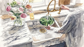 Manos sujetan un colador con judías verdes bajo un grifo que vierte agua en un fregadero de cocina, junto a un florero con flores y una botella.