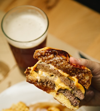 Primer plano de una mano sosteniendo una hamburguesa a medio morder, mostrando capas de queso derretido, carne y un pan tostado. Una cerveza está borrosa al fondo