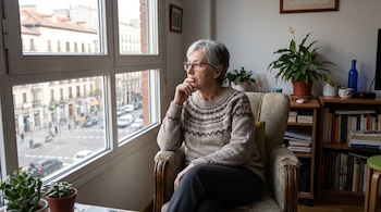 Una mujer con gafas y jersey sentada en un sillón junto a una ventana, con plantas y una estantería con libros alrededor.