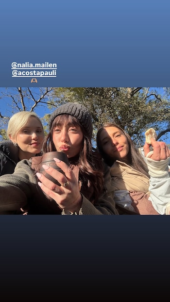 Tres mujeres posan al aire libre. La mujer del centro viste gorro de lana y sostiene un mate. La mujer de la derecha sostiene un trozo de pan