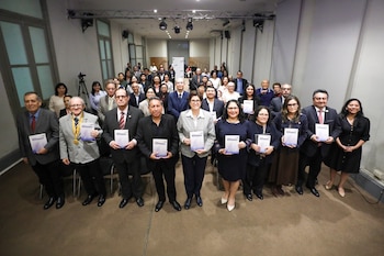 Un grupo de personas sonríe y sostiene ejemplares de la revista NOMBRES. Detrás se observa a una audiencia sentada en un auditorio
