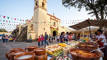 Jarros de barro, elotes preparados y cazuelas sobre una mesa frente a una iglesia adornada con banderines de colores y visitantes.