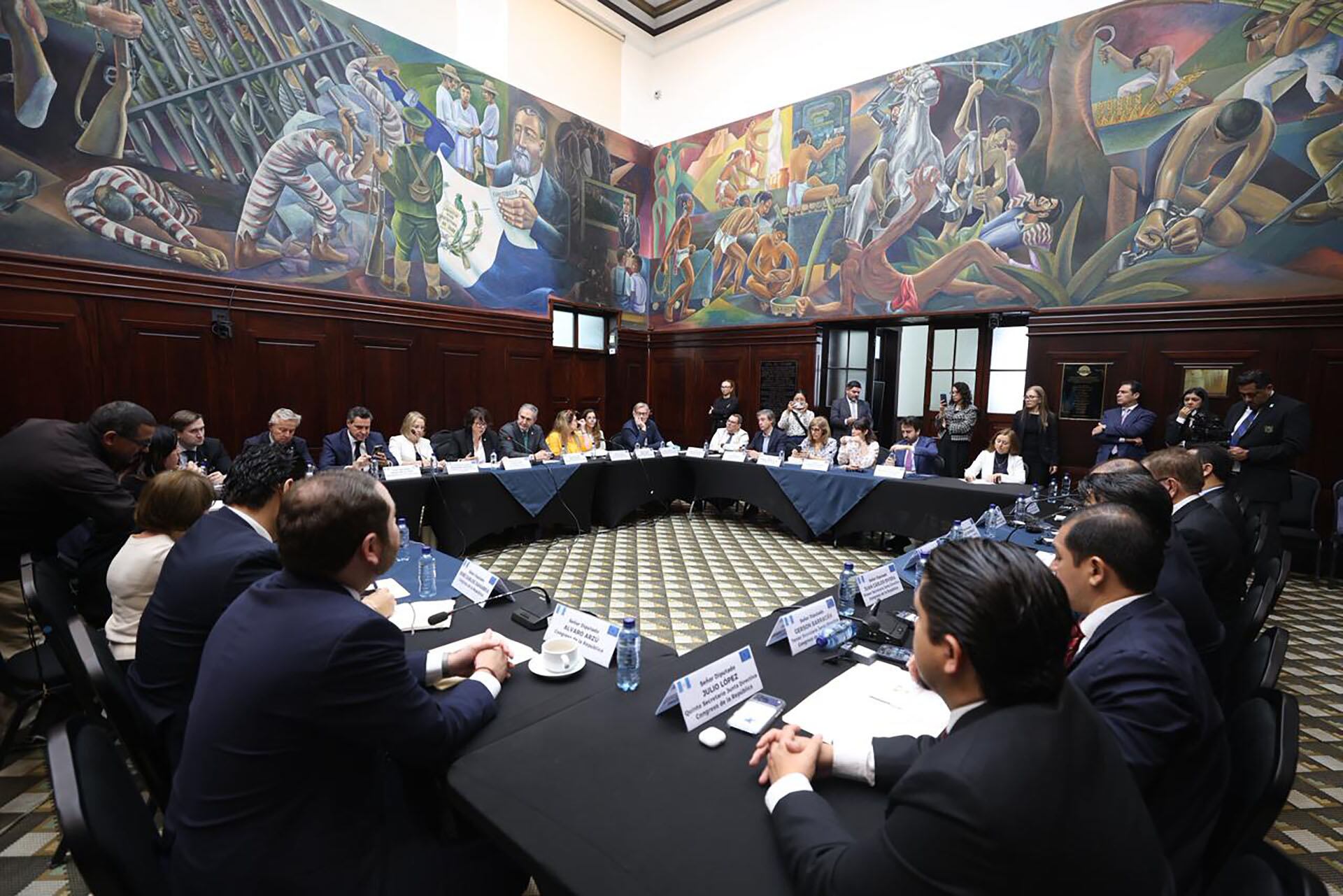 Diversos delegados y líderes participan en una convención formal, sentados alrededor de mesas en forma de U en una sala adornada con grandes murales históricos que narran pasajes importantes. (Organismo Legislativo)