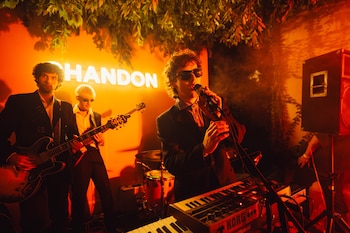 Un músico con gafas de sol canta en un micrófono frente a un teclado, mientras un guitarrista toca detrás, bajo luces naranjas y follaje