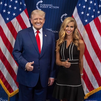 Donald Trump y Katie Zacharia levantan el pulgar ante un fondo con el logotipo de Turning Point Action y dos banderas de Estados Unidos