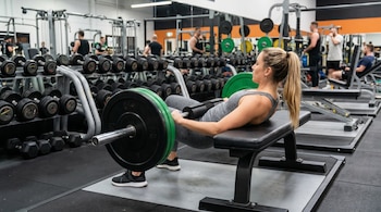 Mujer rubia con cola de caballo en ropa deportiva gris realizando hip thrust con barra y discos verdes en un banco de gimnasio.