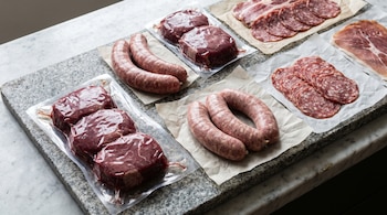 Medallones de carne envasados al vacío, salchichas frescas y lonchas de embutido dispuestas sobre papel y una placa de granito.